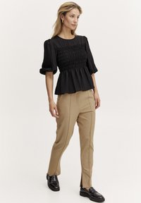 Blusa nera arricciata con maniche a sbuffo corte, abbinata a pantaloni beige su misura e mocassini neri. Tessuto liscio con una leggera texture.