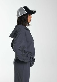 Sudadera con capucha gris con bordado decorativo negro en la manga, combinada con una gorra de malla en blanco y negro. Se muestra en vista de perfil.