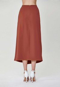 Jupe midi de couleur rouille avec une texture lisse, fermeture éclair au dos et plis subtils. Portée avec des talons hauts blancs à lanières.