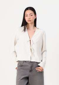 Hvit bluse med lange ermer, vertikale svarte striper, V-hals og knyting foran. Stoffet ser teksturert og lett ut.