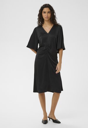 Naine, kes seisab mustas V-kaelusega midi-kleidis, millel on voltis esiosa ja laiad küünarvarre pikkusega varrukad, kandmas musti tipp-trotlitega madala kontsaga sandaale.