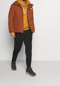 Pánská hnědá parka s kapucí, zapínáním na zip, bočními kapsami, vrstvená přes žlutou mikinu s kapucí, v kombinaci s černými kalhotami a turistickými botami.