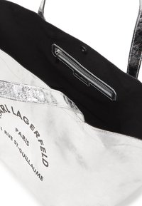 Sac fourre-tout argenté avec une finition métallique lisse, doublure en coton noir, poche zippée à l'intérieur et logo imprimé sur un côté.