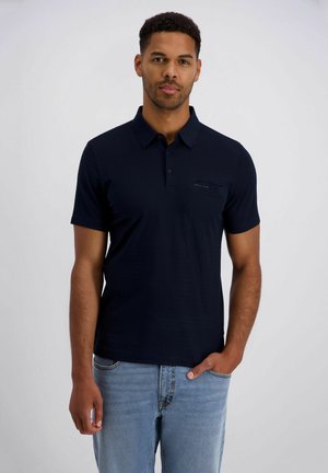 Mann mit kurzen lockigen Haaren, der ein dunkelblaues Polohemd und hellblaue Jeans trägt, vor einem schlichten hellen Hintergrund stehend.