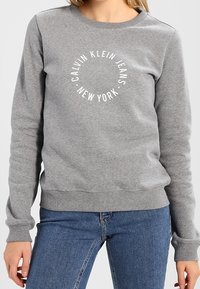 Felpa grigia in misto cotone, con scollo rotondo e polsini a costine. Il testo stampato sul fronte recita "CALVIN KLEIN JEANS NEW YORK."