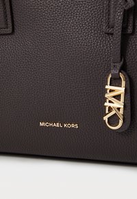 Gros plan sur un sac en cuir brun foncé texturé avec le logo doré Michael Kors et un porte-clés doré MK attaché à la bandoulière.