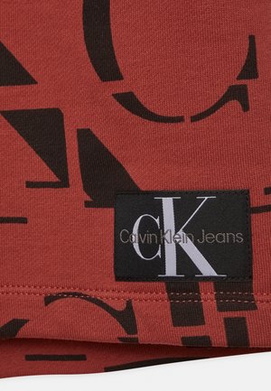 Zwarte Calvin Klein Jeans logo tag op rood stof met grote zwarte abstracte letters en zichtbare stiksels.