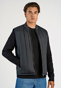 Vadderad svart väst med en ribbad svart tröja under. Vesten har dragkedja fram och sidfickor med dragkedja, samt vertikala quiltade linjer.