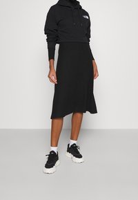 Černá krátká mikina s kapucí s grafickým logem, doplněná černou sukní po kolena. Nošeno s černými teniskami s tlustou bílou podrážkou.