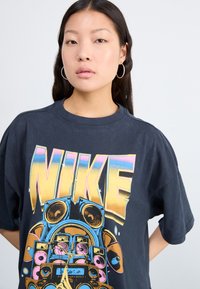 Črna bombažna majica s kratkimi rokavi, ki prikazuje velik večbarvni grafični napis "NIKE" z retro zvočniki. Okrogla izreza in sproščen kroj. Gladka tekstura.