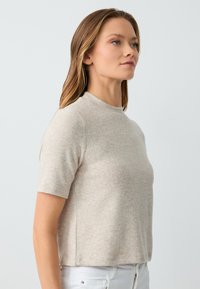 Top beige a maniche corte realizzato in tessuto morbido e testurizzato. Presenta un collo alto e una silhouette aderente, abbinato a pantaloni bianchi.