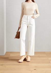 Weiße hoch taillierte weite Jeans, kombiniert mit einem beige langen Oberteil und beige Blockabsatzstiefeln. Braune Handtasche mit einem kurzen Tragegurt.
