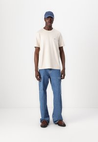 T-shirt en coton beige à manches courtes, pantalon cargo en denim avec poches latérales, et casquette bleue, se tenant sur un fond neutre.