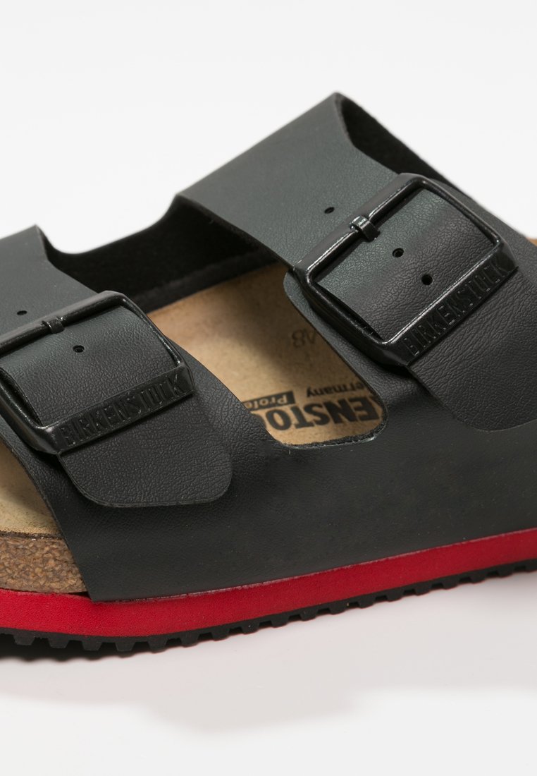 Black leather sandal with two adjustable straps, a cork footbed, and a contrasting red rubber sole. Features signature branding on the strap.