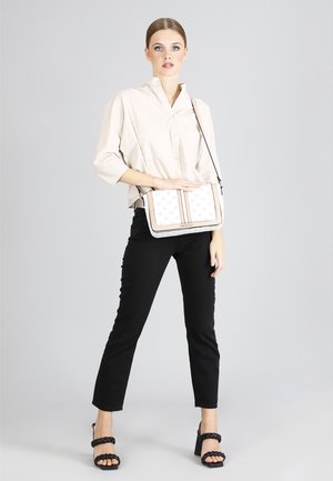 Borsa a spalla bianca con dettagli beige e un design con motivo. Modella che indossa una camicia beige, pantaloni neri e tacchi neri con cinturini.
