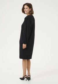 Abito in maglia nera con maniche lunghe e plissettate, silhouette dritta, lunghezza al ginocchio, abbinato a scarpe con punta a stiletto nere. Design semplice ed elegante.