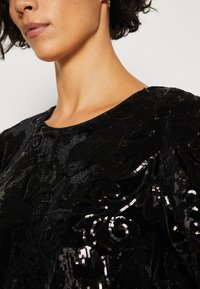 Robe noire avec une surface texturée, ornée de motifs en sequins noirs délicats et d'un col rond. Tissu lisse avec un léger éclat.