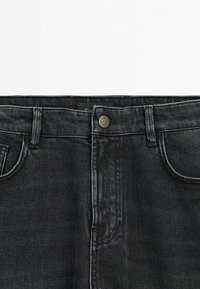 Ceinture de jeans en denim noir avec fermeture par bouton, passants pour ceinture et poches avant visibles sur un fond clair uni.