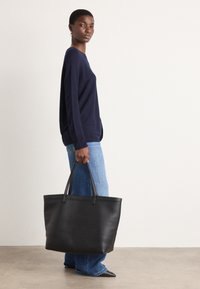 Svart lær tote-veske med dobbelte håndtak, rektangulær form, glatt tekstur, brukt sammen med en marineblå genser og blå jeans.