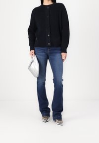 Cardigan de punto azul marino con cuello redondo y cierre de botones, combinado con jeans acampanados azules y un bolso de mano plateado.