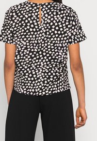 Persona vestida con una blusa negra de manga corta con patrones cuadrados irregulares blancos y pantalones negros, vista desde la espalda contra un fondo liso.
