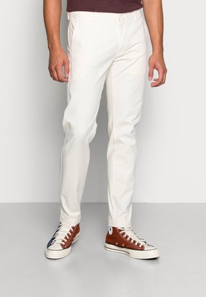 Homme portant un pantalon blanc slim et des baskets montantes bicolores, debout devant un fond clair uni.