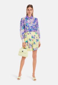 Lila Blumenbluse mit hohem Kragen, kombiniert mit einem gelben Rock, der mit blauen und lila Blumen verziert ist. Als Accessoires dienen eine gelbe Handtasche und Sandalen.