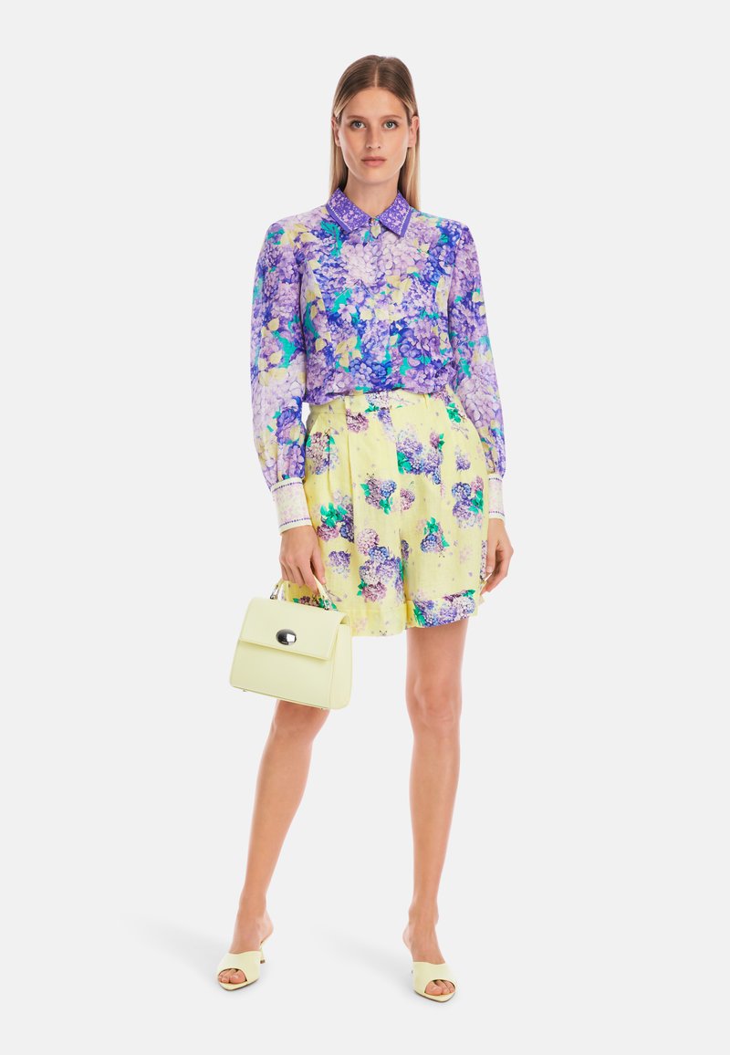 Lila Blumenbluse mit hohem Kragen, kombiniert mit einem gelben Rock, der mit blauen und lila Blumen verziert ist. Als Accessoires dienen eine gelbe Handtasche und Sandalen.