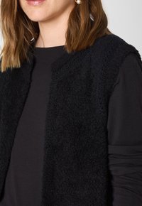Gilet nero, strutturato, realizzato in un materiale morbido e peloso, senza chiusure, indossato sopra una maglietta nera a maniche lunghe semplice.