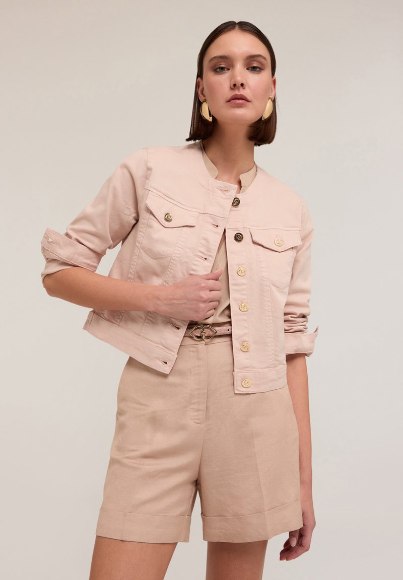Ljusrosa, croppad denimjacka med två bröstfickor och gyllene knappar, kombinerad med beiga shorts och ett matchande bälte med en gyllene spänne.