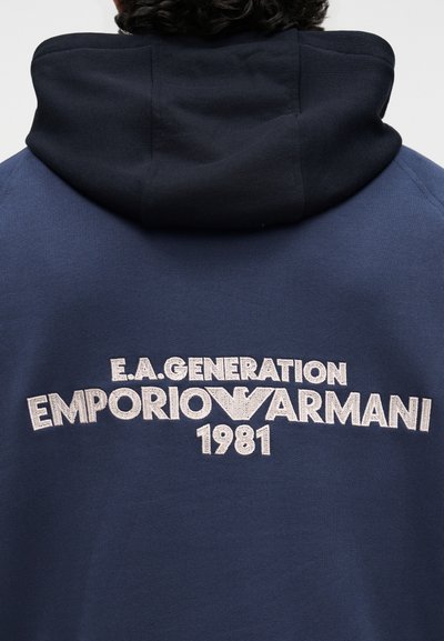 Személy sötétkék kapucnis pulcsiban, fekete kapucnival, amelyen fehér hímzett "E.A. Generation Emporio Armani 1981" felirat található a háton.