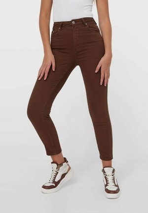 Persoon draagt bruine high-waist skinny jeans en witte-bonte hoge sneakers, staat met gespreide benen en handen rustend op de dijen.