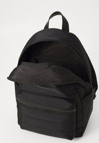 Sac à dos noir avec une poignée supérieure, compartiment principal ouvert montrant une poche zippée intérieure, et poche zippée avant sur fond uni.