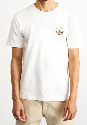 T-shirt en coton blanc à manches courtes, col rond, présentant un petit logo Adidas violet et or sur le côté gauche de la poitrine.