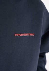Tmavomodrý textil so slovom "ZAKÁZANÉ" vyšitým červenou farbou, umiestneným na strednej časti hrudníka odevu.