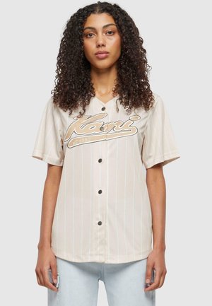 Helles beige Kurzarm-Jersey mit vertikalen Streifen, Knopfleiste vorne und "Kani"-Logo in Schriftzug über der Brust, verziert mit schwarzen Knöpfen.