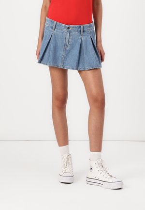 Persoon draagt een geplooide lichtblauwe denim minirok, witte enkelsokken, witte hoge sneakers en een rood topje, staand op een witte achtergrond.