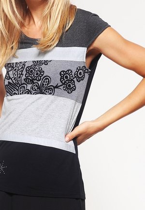 Femme portant une chemise à manches courtes avec des panneaux horizontaux gris et noirs, ornée d'un imprimé floral sur la poitrine, la main posée sur la hanche.