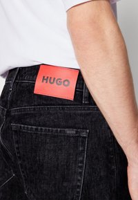 Gros plan d'une personne portant un jean en denim noir avec une étiquette rouge de la marque HUGO à la taille et une chemise blanche rentrée.