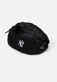 Pochette zippée en polaire noire avec logo blanc "NY" sur le devant, partiellement ouverte montrant l'intérieur en tissu noir.
