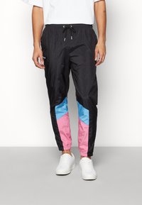 Man wearing black track pants with blue and pink color blocks near ankles and white sneakers, standing against white background.