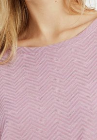 Nainen pukeutuneena liilanväriseen neulepaitaan, jossa on siksak-kuvio ja venemäinen kaula-aukko, joka paljastaa osan hänen kaulaansa ja vaaleita hiuksiaan.