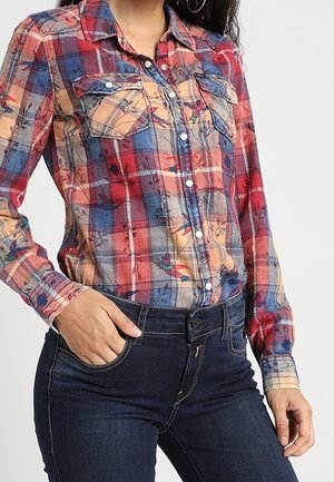 Rot-blau kariertes Hemd mit Knöpfen, floralen Akzenten, zwei Brusttaschen und einem Kragen. Getragen mit dunkelblauen Jeans.