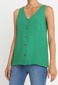 Grön, ärmlös topp med V-ringning och knäppning fram. Tillverkad av krinklad tyg, med en avslappnad passform och sömmar.