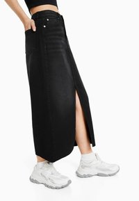 Černá maxi džínová sukně s rozparkem vpředu a bočními kapsami, s vysokým pasem a olemovaným lemem. Kombinována s bílými atletickými teniskami.