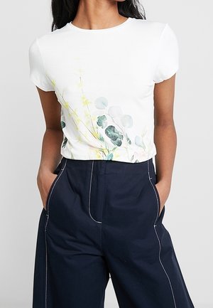 Kobieta ubrana w biały top z krótkim rękawem z delikatnym zielonym kwiatowym nadrukiem, włożony do wysokich, granatowych spodni z białymi przeszyciami.