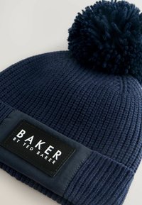 Navy gebreide beanie met een grote pompon bovenop en een zwart label met de tekst "BAKER by TED BAKER" op de voorkant gestikt.
