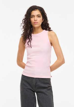 Mujer con pelo rizado, que lleva una camiseta sin mangas rosa claro de canalé y vaqueros gris oscuro, de pie con las manos detrás de la espalda, expresión neutral.