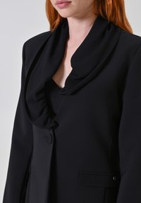 Blazer nero con colletto drappeggiato, texture liscia, chiusura con un solo bottone e tasche laterali, dal taglio sartoriale.