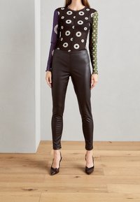 Body nero con motivo floreale, maniche lunghe trasparenti viola e verde, abbinato a leggings in pelle nera e tacchi a punta su un pavimento in legno.